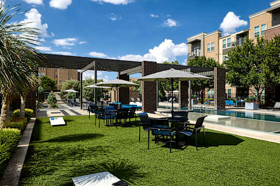 A patio with chairs and umbrellas is surrounded by a grassy area and a pool.