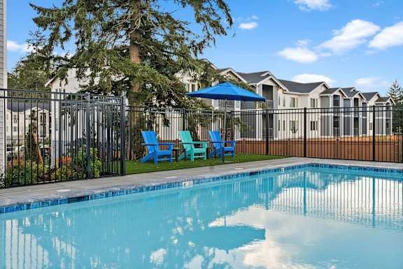 A pool surrounded by a black fence with blue chairs and a tree.
