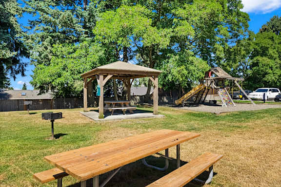 A wooden picnic table is in the foreground of a park with a gazebo and playground in the background.