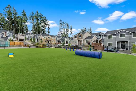 A large grassy field with a blue cylinder in the middle and houses in the background.