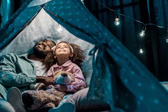 A man and a little girl are lying in a tent with a teddy bear.