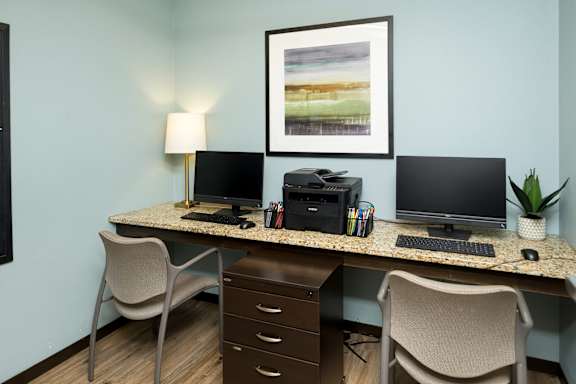 A desk with two monitors, a keyboard, a mouse, a lamp, a plant, and a framed picture on the wall.