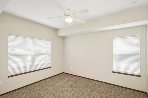 A room with a carpeted floor, a ceiling fan, and two windows with blinds.