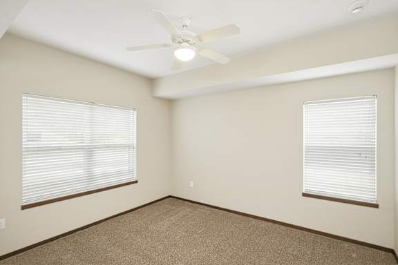 A room with a carpeted floor, a ceiling fan, and two windows with blinds.