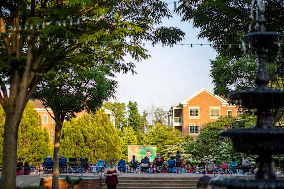 A fountain is in the foreground of a park with people sitting on chairs in the background.