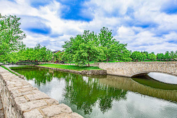 A stone wall borders a body of water with a bridge crossing over it at French District Villas Apartments, St. Charles, 63301