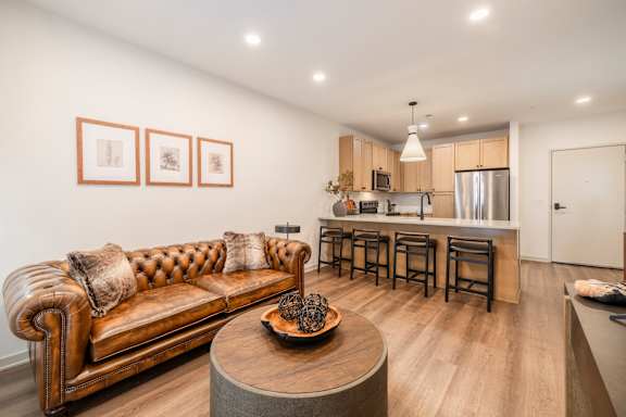 A brown leather couch with pillows sits in a living room with a kitchen in the background.