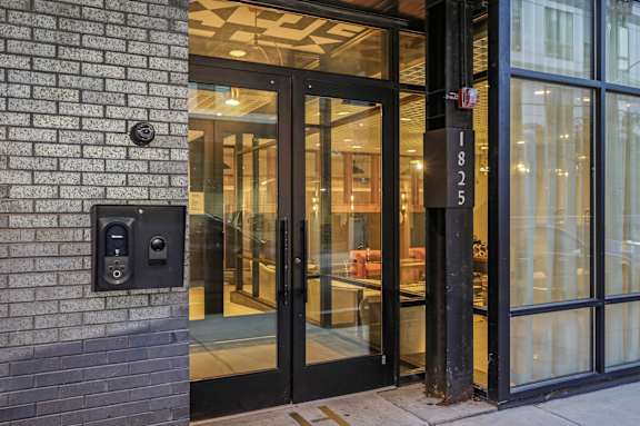 The image shows a modern building entrance with a glass door and a brick wall on the left side.
