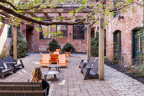 A woman is sitting on a bench under a pergola in a courtyard with brick walls and trees.
