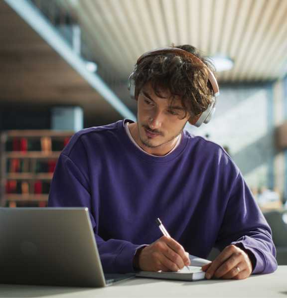 A person in a purple sweater is writing on a notepad while using a laptop.