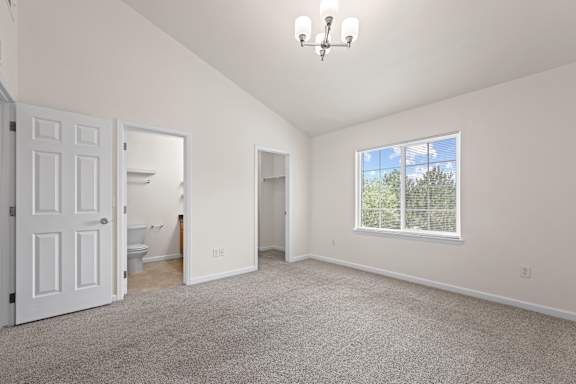 A large, empty room with a carpeted floor and a window showing a view of trees outside.