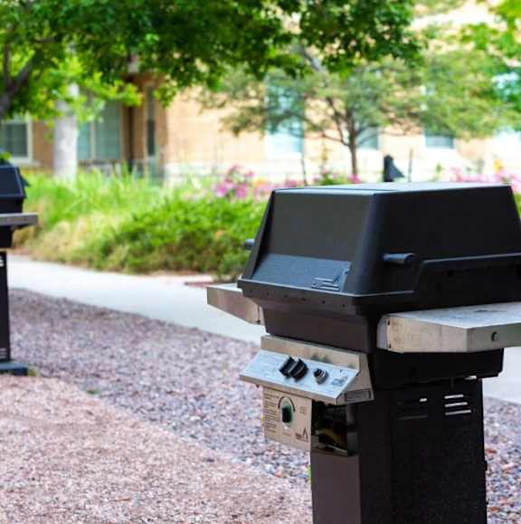 A black parking meter is on a gravel area with trees and a building in the background.