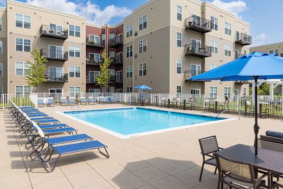 A pool surrounded by chairs and umbrellas in front of apartment buildings.