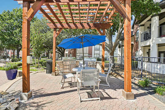 A wooden pergola with a blue umbrella is in the foreground of a brick patio.