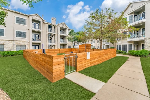 A wooden fence with a gate is in the foreground of a grassy area with apartment buildings in the background.