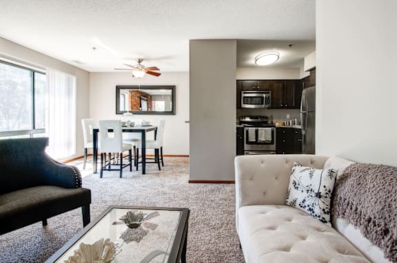 A modern living room with a black couch, a glass coffee table, and a kitchen in the background.