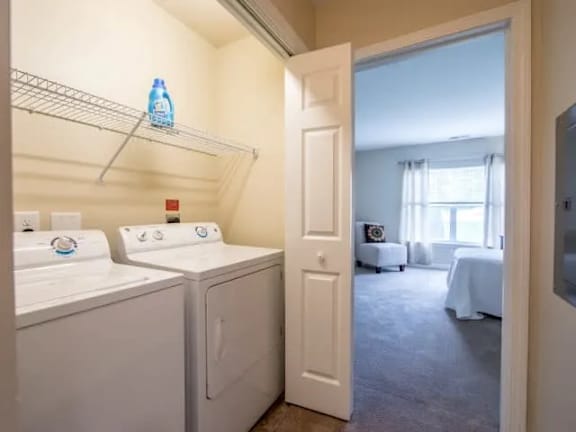 A laundry room with a washer and dryer, a shelf with a water bottle, and a view into a living room.