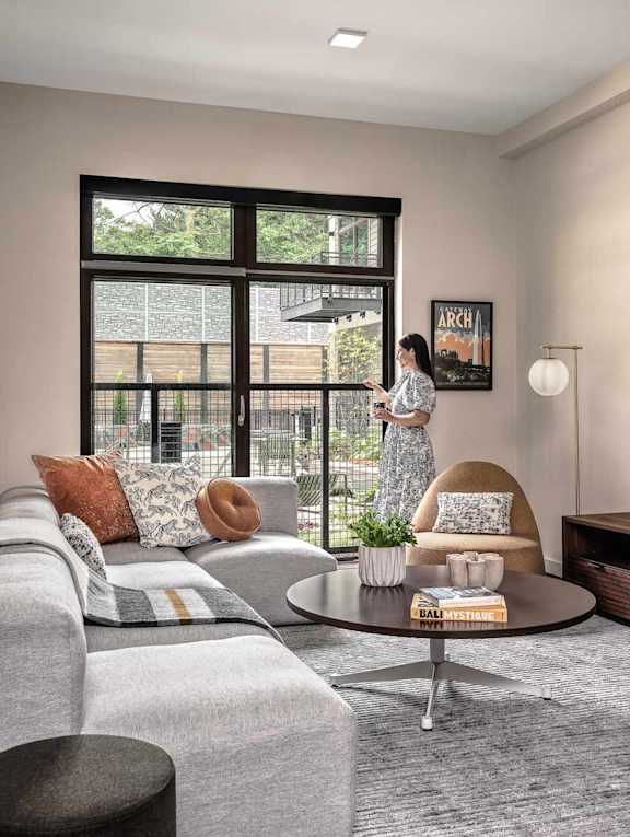 A woman stands in a living room with a grey couch and a round table.