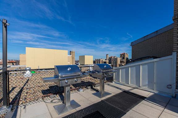 A grill is on a patio with a white fence and a building in the background.