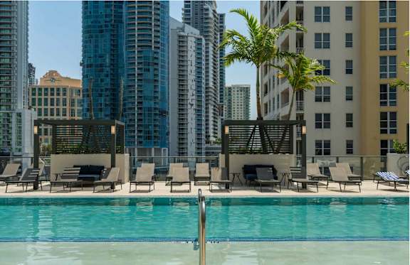 A pool surrounded by chairs and palm trees with a city skyline in the background.