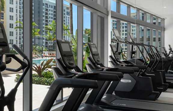 A row of treadmills in a gym with a view of a pool and buildings outside the window.
