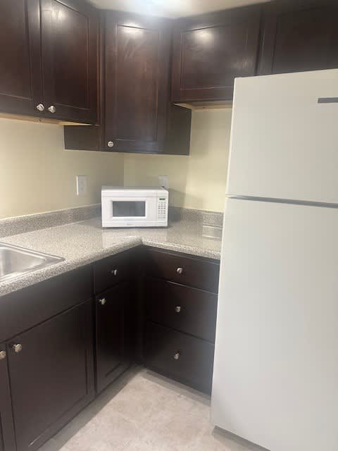 A white refrigerator stands next to a microwave in a kitchen with dark brown cabinets.