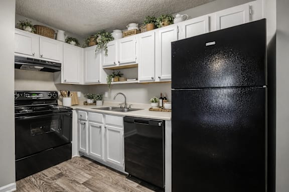 A black fridge stands in a kitchen with white cabinets and a wooden floor.