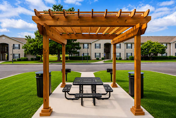 A wooden pergola with a table and chairs is in the foreground of a grassy area with apartment buildings in the background.