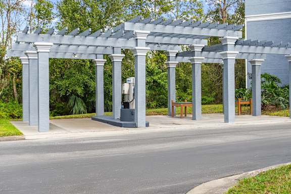 A grey pergola with columns and benches is situated on a paved car wash area.