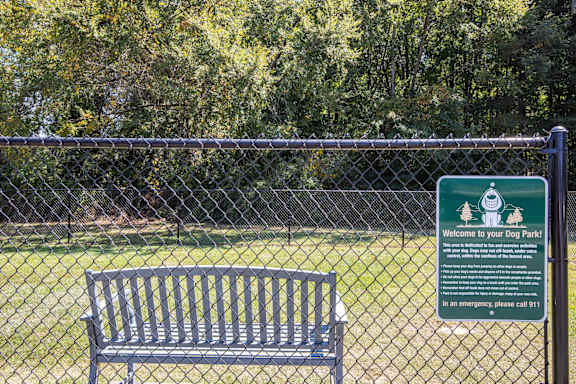 A sign welcomes visitors to a dog park with a bench in front of a fence.