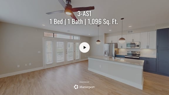 A spacious room with a fan and a kitchen area in the background.