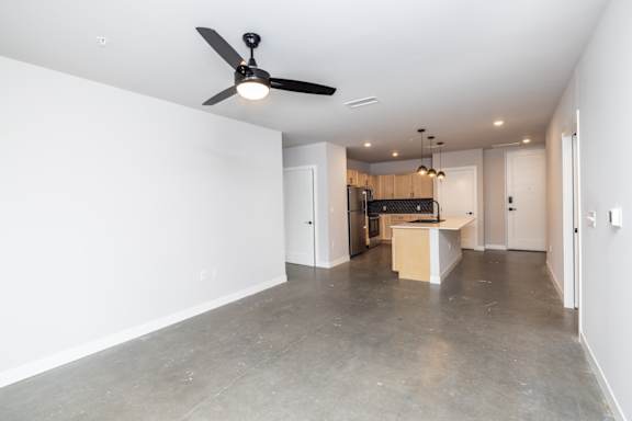 A spacious empty room with a ceiling fan and a kitchen area in the background.