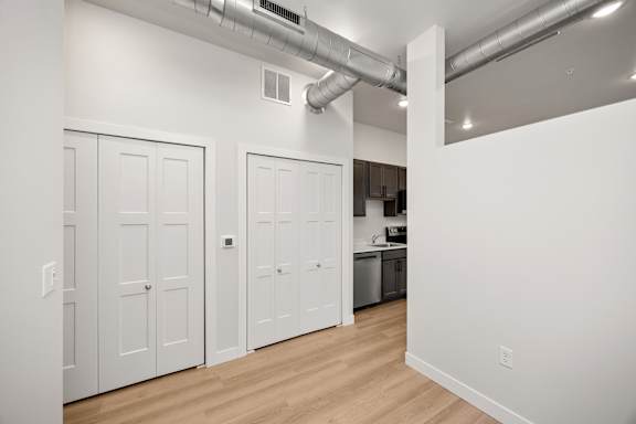 A room with white walls and wooden floors, with a kitchen area visible in the background.