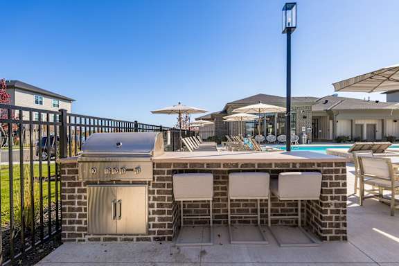 A brick barbecue grill with two chairs and an umbrella in front of a pool.