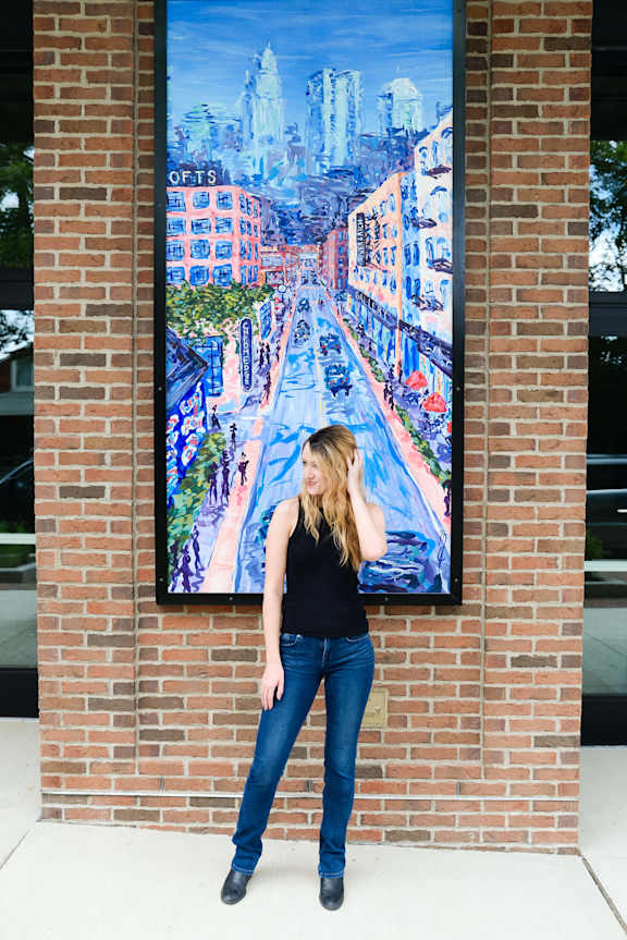 A woman poses in front of a large mural of a city street.