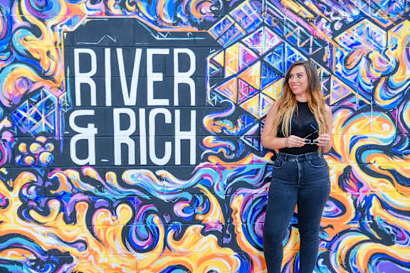 A woman stands in front of a colorful wall with the words "River & Rich" written on it.