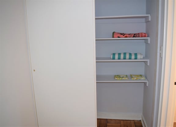 A white closet with three shelves, the top shelf has a pink and black striped towel, the middle shelf has a green and white striped towel, and the bottom shelf has two yellow towels