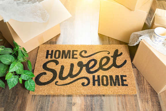 A doormat with the words "Home Sweet Home" on it is in the foreground with boxes and a plant in the background.