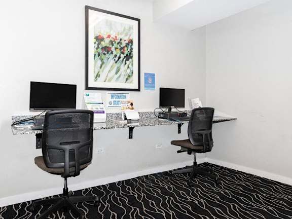 Two black chairs are in front of a desk with a computer monitor and a framed picture on the wall.