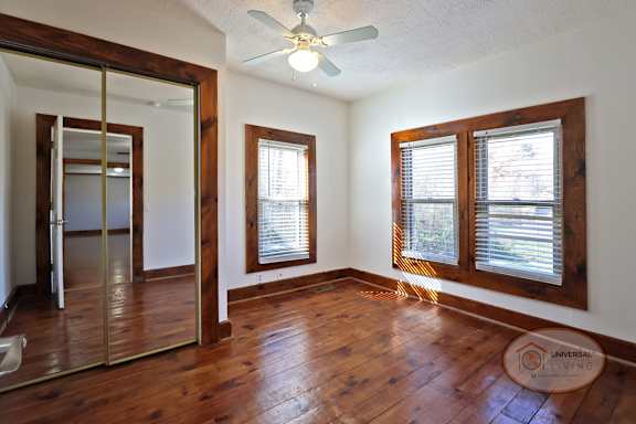 A bedroom with hardwood flooring, large windows, ceiling fan, and mirrored closet door.