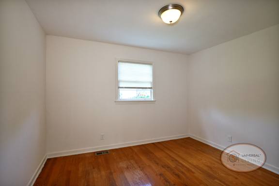 A bedroom with white walls, hardwood flooring, and a window.
