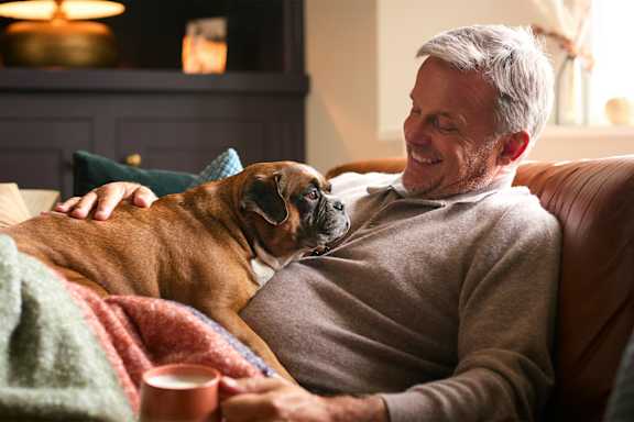 A man is sitting on a couch with a dog and a cup of coffee.