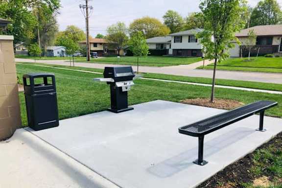A black trash can and a black bench are on a concrete slab in a grassy area.