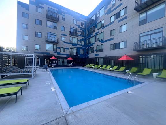 A large swimming pool surrounded by lounge chairs and umbrellas in front of a multi-story apartment building.