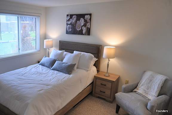 A bedroom with a large bed, a nightstand with a lamp, and a chair with a blanket on it.