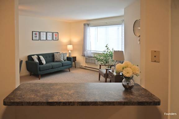 A living room with a dark green couch, a coffee table with a vase of flowers, and a window with white curtains.
