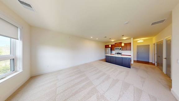 A large, empty room with a carpeted floor and a kitchenette in the background.