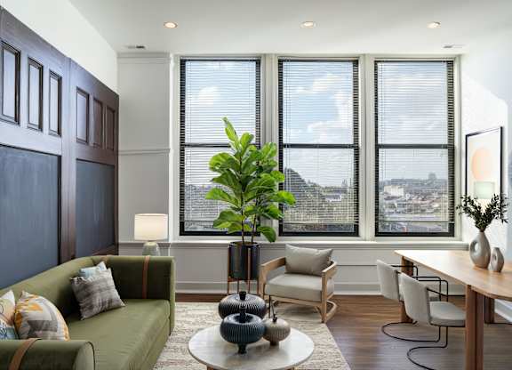 A living room with a green couch, a table with a vase and a potted plant, and a view of the city through the windows.