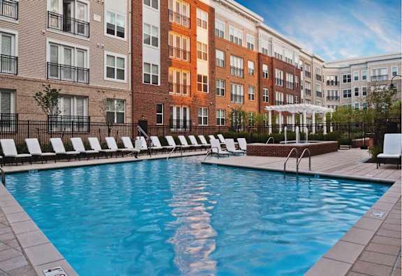 A swimming pool surrounded by lounge chairs in front of a row of apartment buildings.