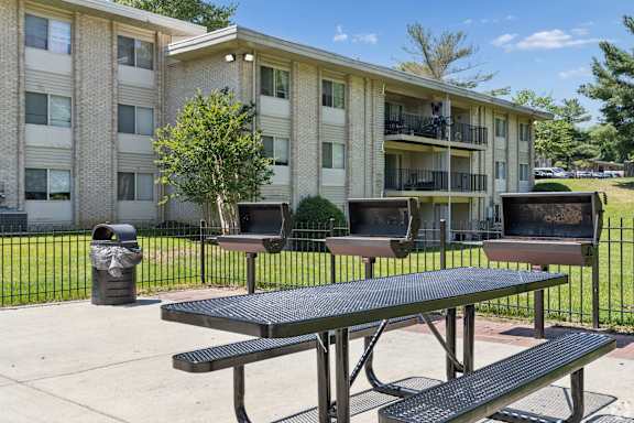 A black picnic table is in front of a building with a tree and a trash can in the foreground.